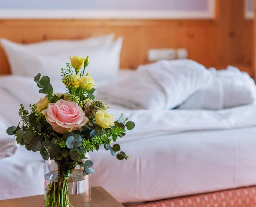 kleiner Blumenstrauß mit einer rosa Rose und kleinen gelben Blüten im Vordergrund, hinten ein Bett mit zwei Bademänteln Valentinswochenende am Brombachsee